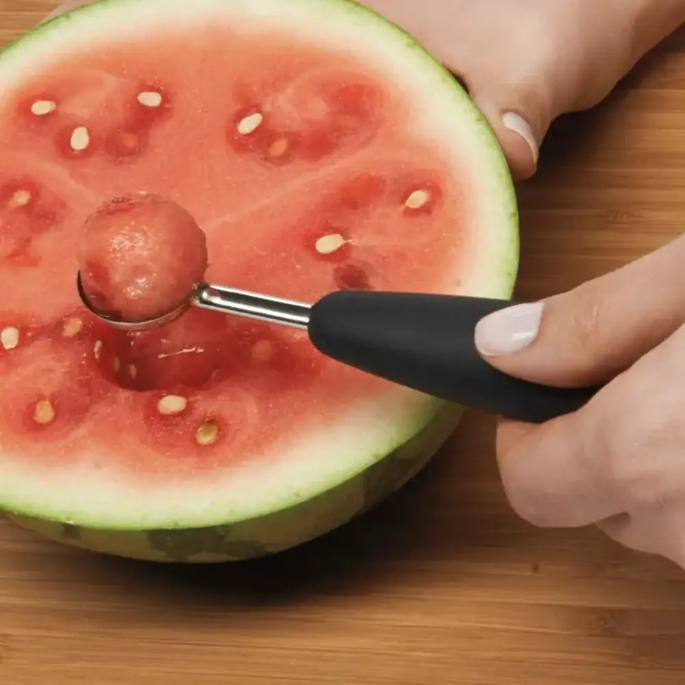 Person using a watermelon baller to scoop out a piece of watermelon on a wooden surface.