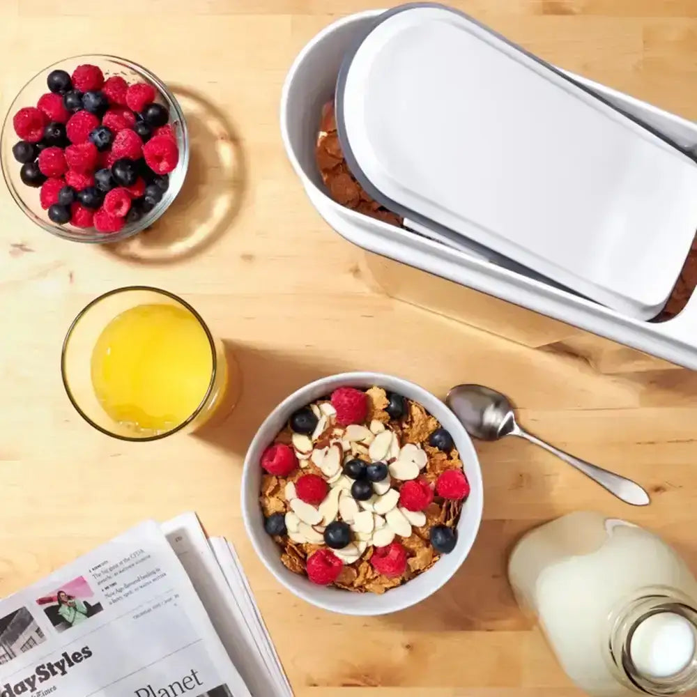 Breakfast setup with cereal bowl, juice, berries, and newspaper on a wooden table.