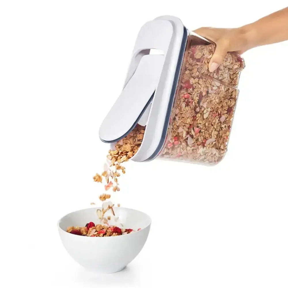 Hand pouring granola from a transparent container with blue lid into a white bowl on a white background