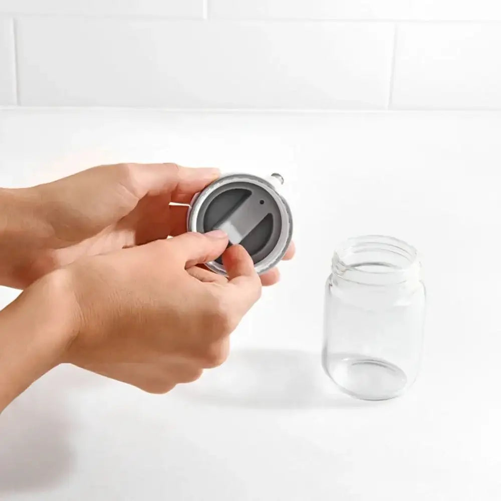 Person holding a silver lid over a clear glass jar on a white background