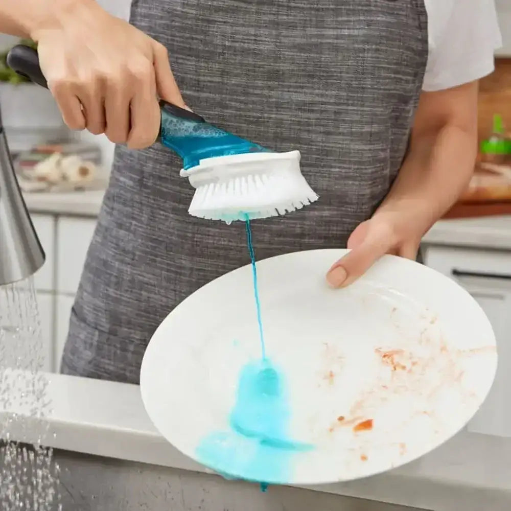 Person using a blue scrubber to clean a white plate with food residue in a kitchen.