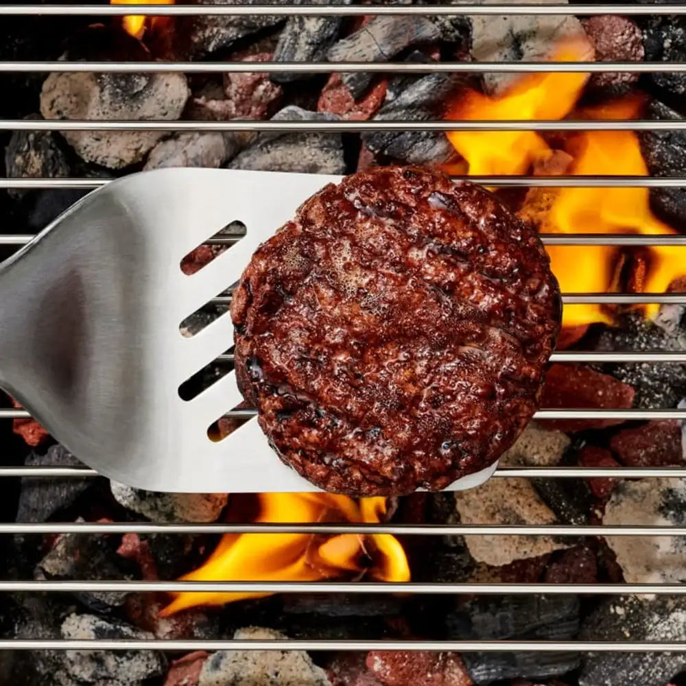 Hamburger patty being flipped on a grill with flames and coals visible