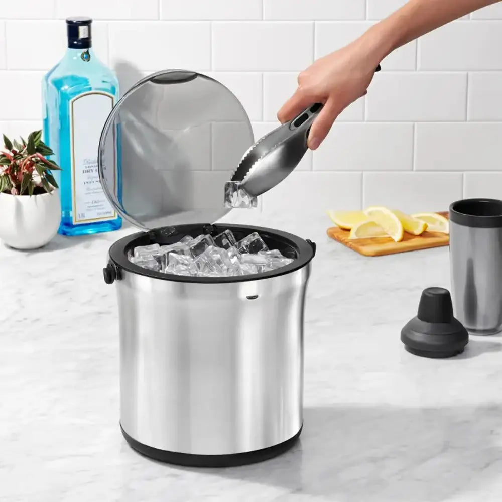 Stainless steel ice bucket with a hand using tongs to add ice cubes, set against a kitchen counter.