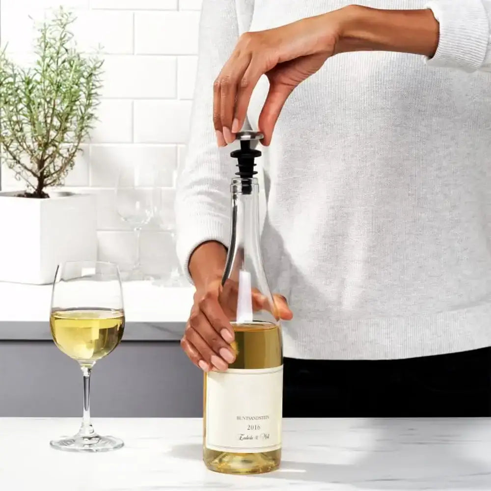 Person opening a bottle of white wine with a wine opener, next to a glass of wine on a kitchen counter.