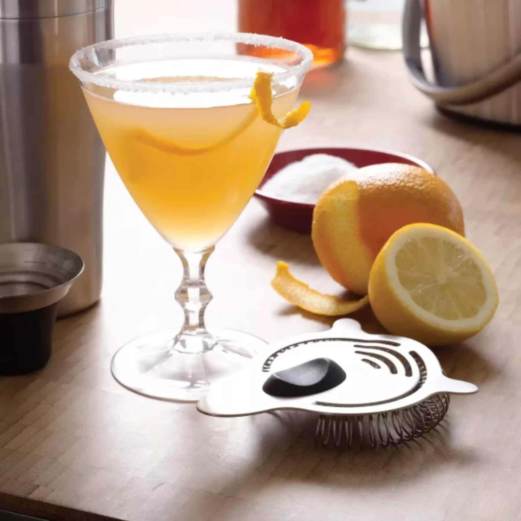 Cocktail glass with a lemon twist on a wooden surface with lemons and a strainer.