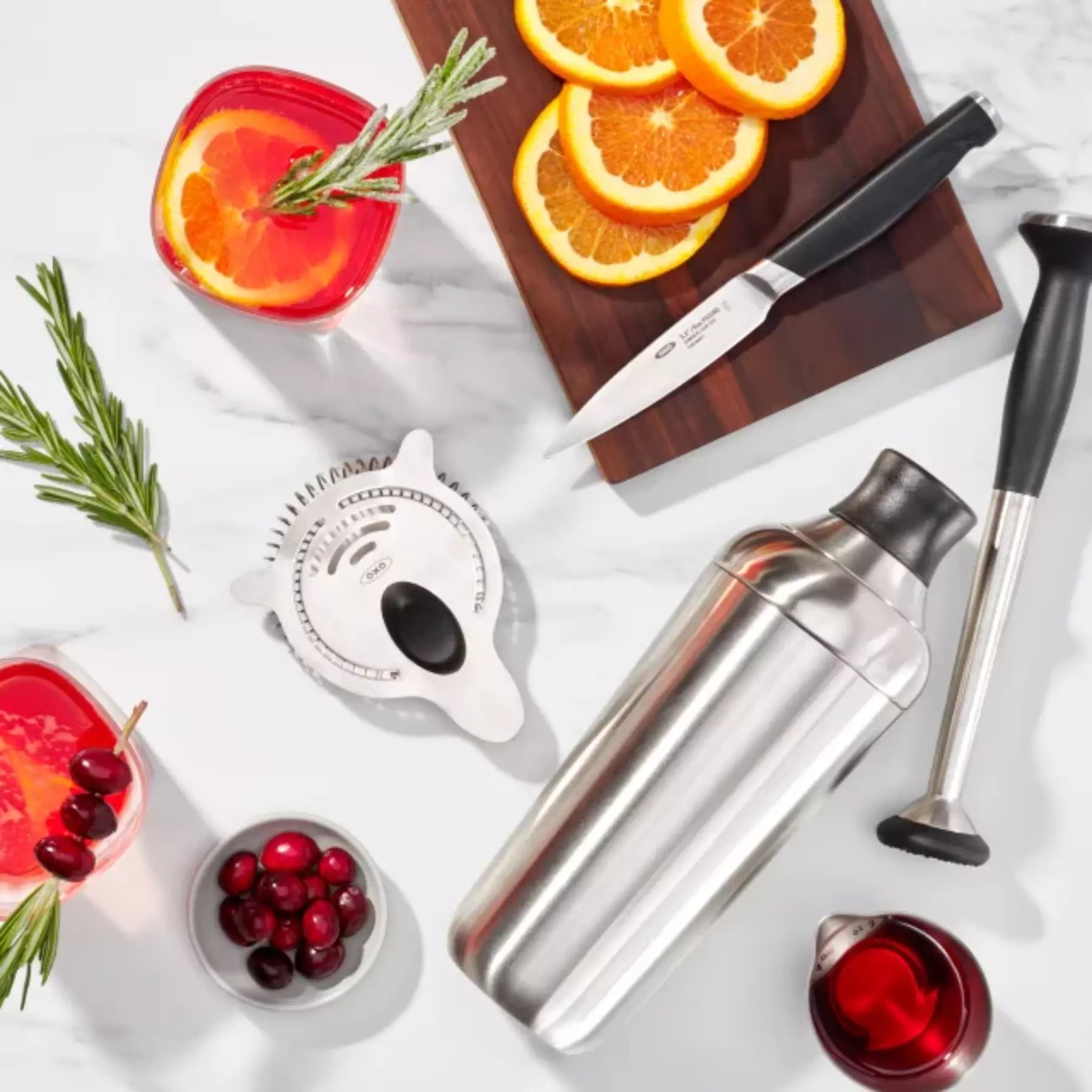 Cocktail-making setup with a shaker, strainer, and ingredients on a white surface.