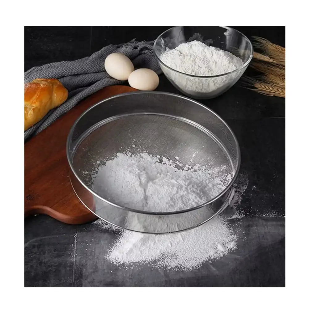 Metal sieve with flour on a dark surface with eggs and bread in the background