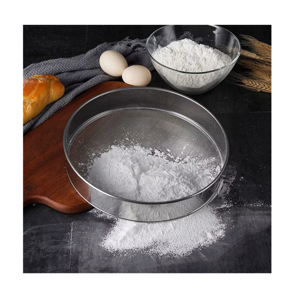 Metal sieve with flour on a dark surface with eggs and bread in the background