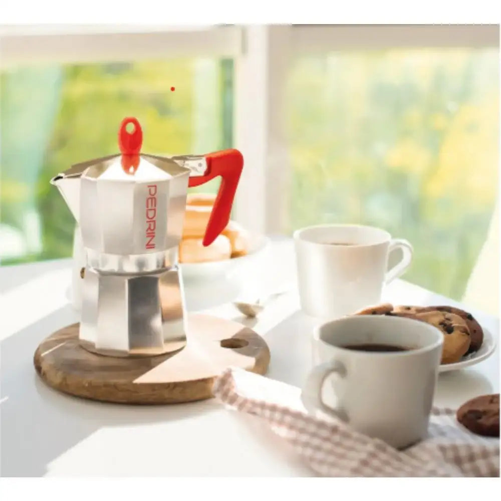 Stainless steel espresso maker on a wooden coaster with two cups of coffee and cookies on a table.