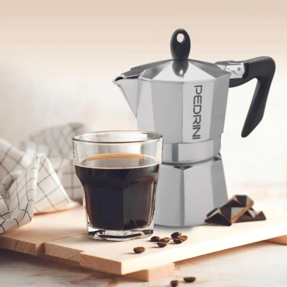 Silver Pedrini coffee maker with a glass of coffee and chocolate on a wooden surface.