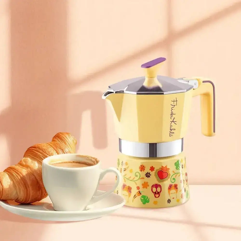 Yellow coffee maker with floral design next to a cup of coffee and croissant on a pink background