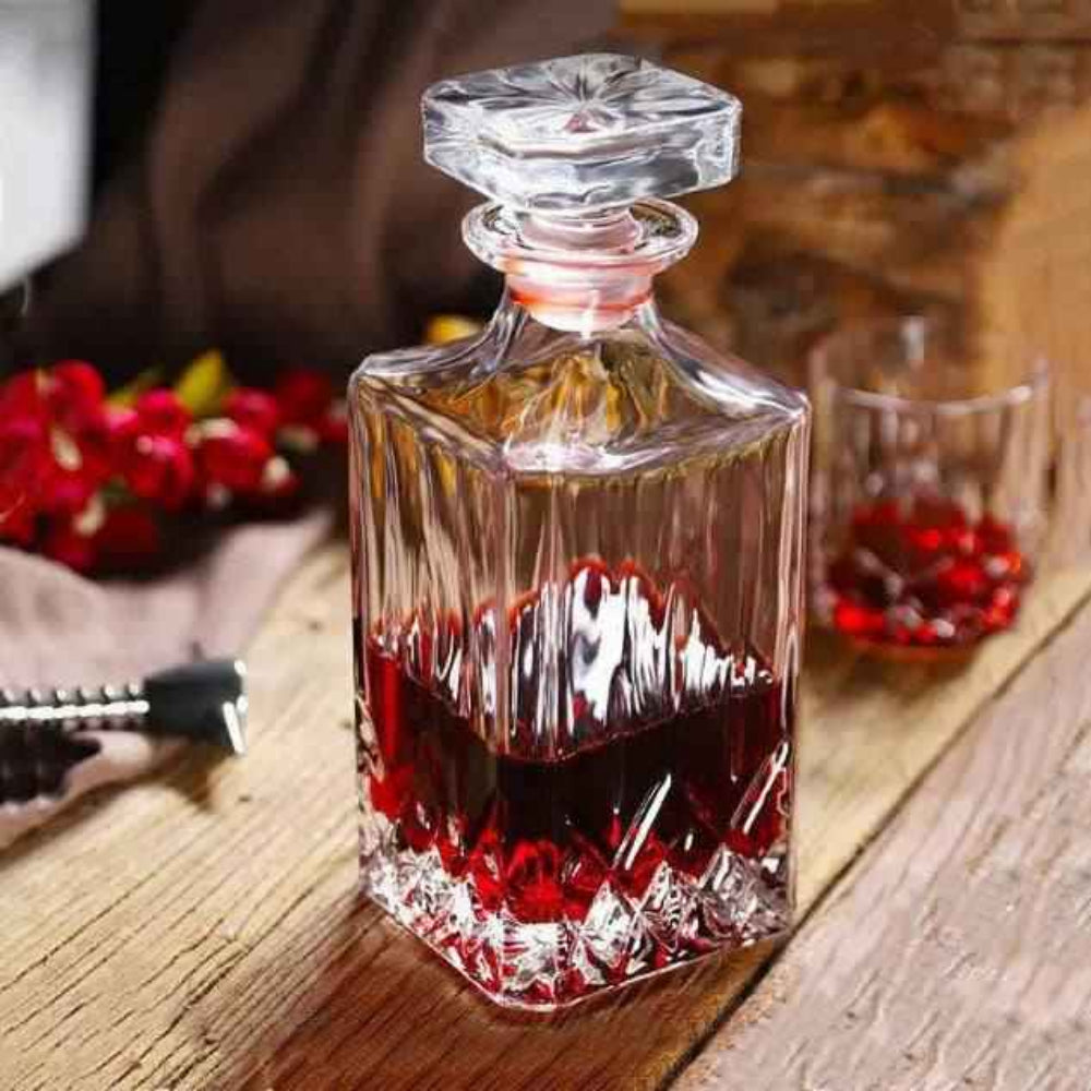 Glass decanter with red liquid on a wooden surface, surrounded by flowers and another glass.