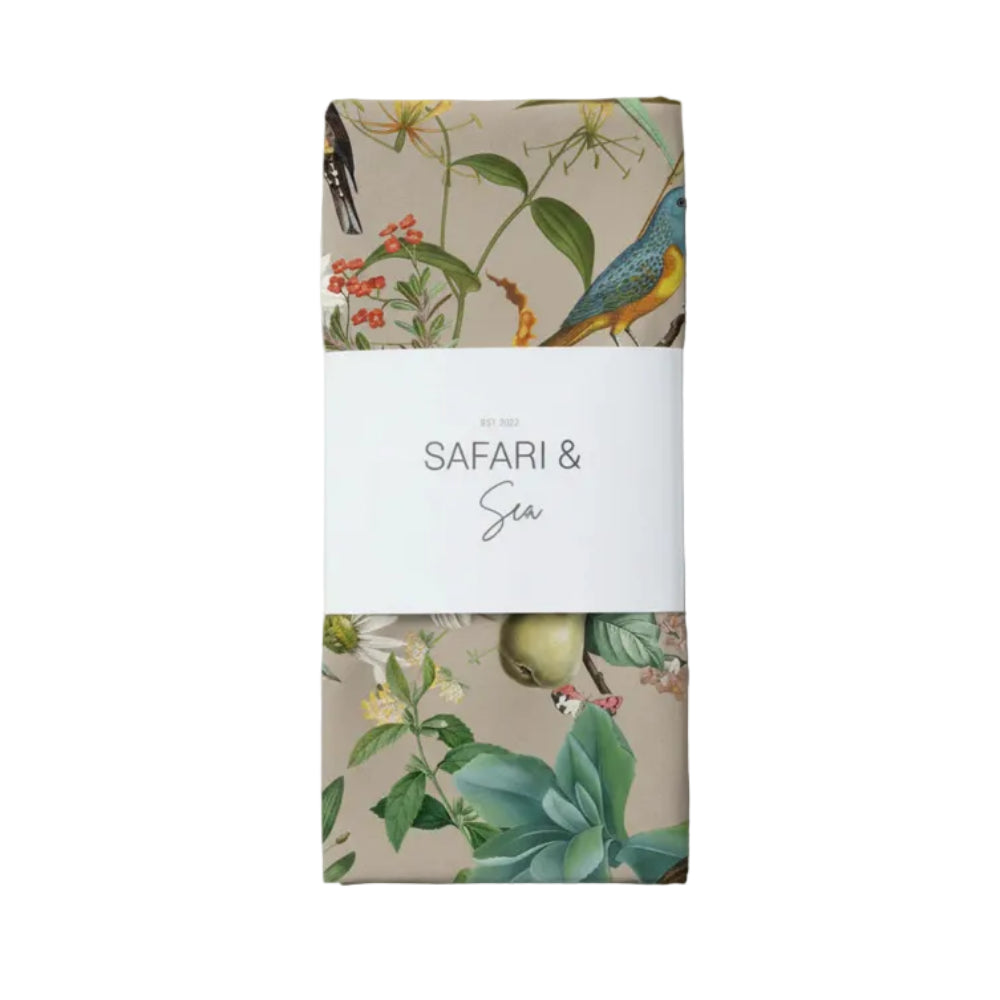 A folded textile product wrapped in a white paper band reading 'SAFARI & Sea', shown on a light gray background. The fabric has a beige safari-style print with illustrated birds, flowers, and green leaves.