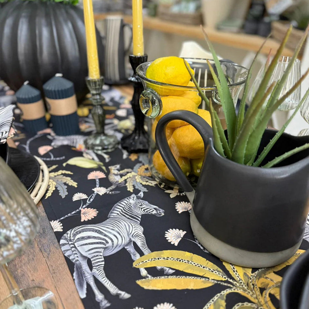 A styled tabletop scene featuring a black table runner with a zebra and botanical print, a dark ceramic pitcher with a succulent, yellow lemons in a glass bowl, and yellow taper candles on a wooden table.