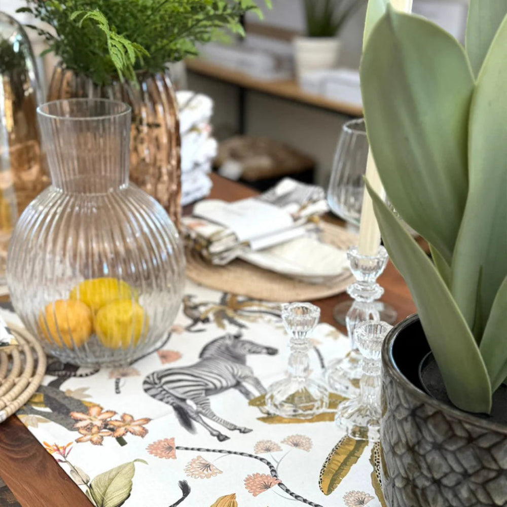 A close-up of a styled dining table featuring a white table runner with a safari print of zebras, leopards, and botanical motifs. The table is set with a ribbed clear glass carafe containing lemons, clear glass candlesticks, white plates, and a large potted green plant in the foreground.