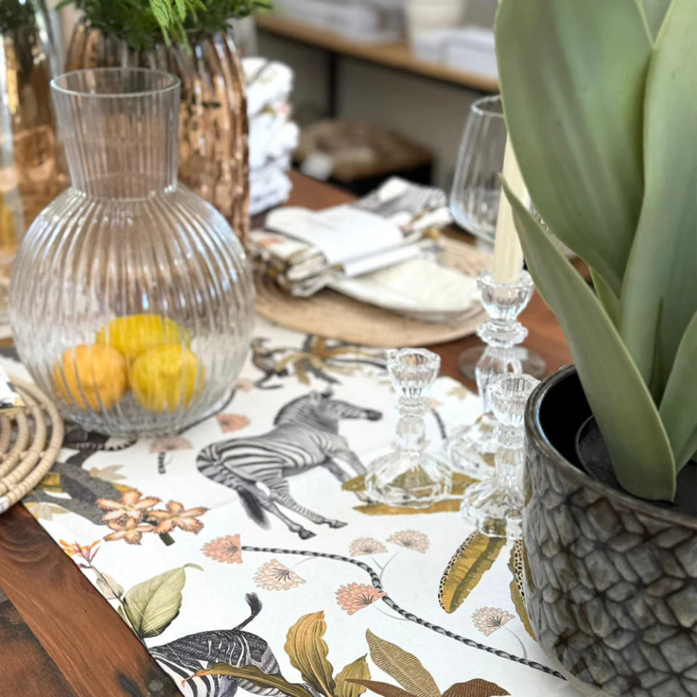 A close-up of a styled dining table featuring a white table runner with a safari print of zebras, leopards, and botanical motifs. The table is set with a ribbed clear glass carafe containing lemons, clear glass candlesticks, white plates, and a large potted green plant in the foreground.