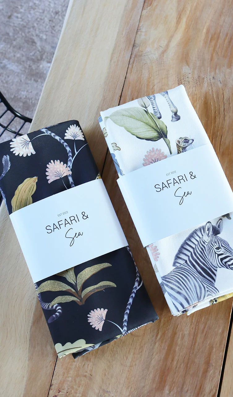 Two folded fabric items wrapped in white paper bands labeled 'SAFARI & Sea' are displayed on a wooden surface. One fabric piece has a dark navy floral print with beige and pink botanical motifs, and the other has a white print featuring a zebra and green leaves.