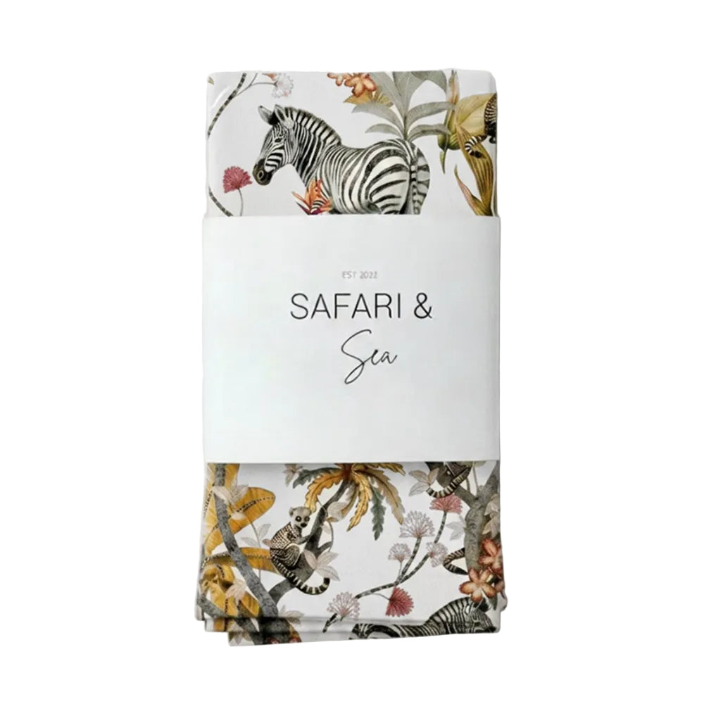 A folded textile product wrapped in a white paper band labeled 'SAFARI & SEA EST. 2022', featuring a safari-themed print with a zebra, palm leaves, and tropical flowers on a light background.