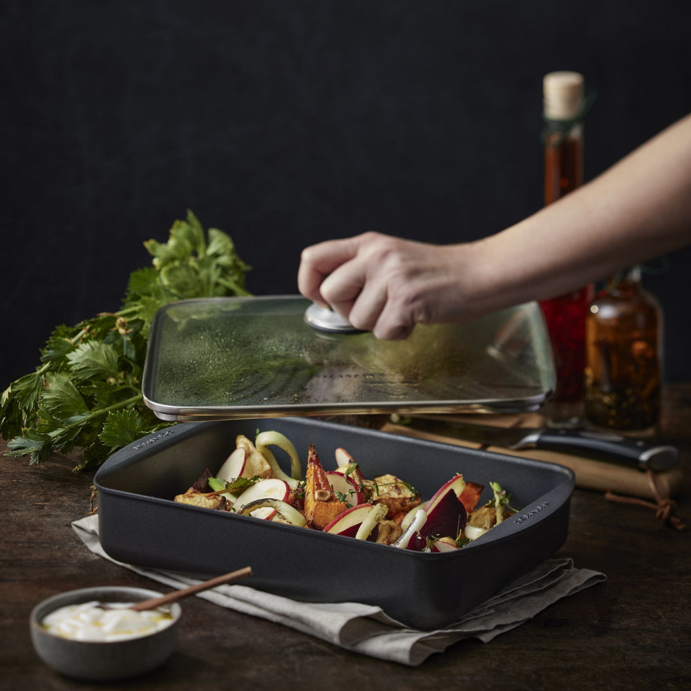A dark gray rectangular cast iron roasting pan filled with roasted vegetables, with a hand lifting a glass lid from a matching roasting pan in a rustic kitchen setting.