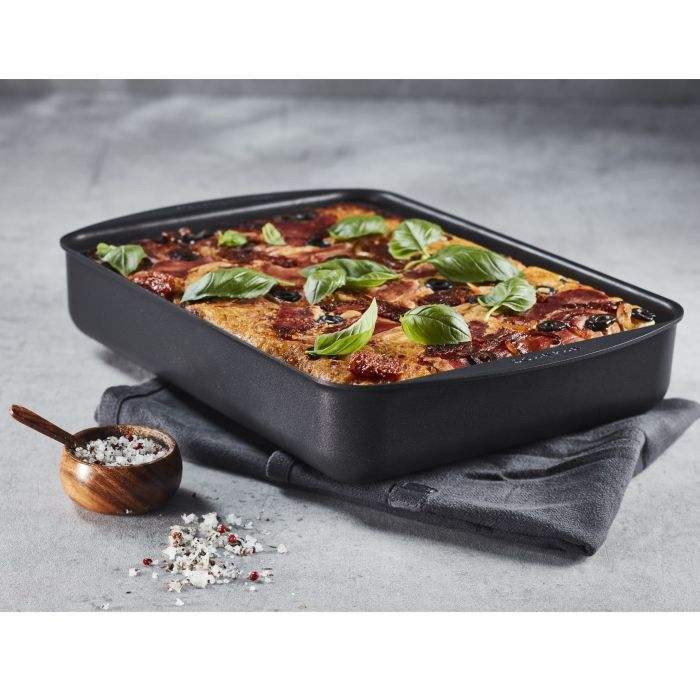 Baked dish in a black rectangular baking tray on a gray surface with a small wooden bowl of herbs.