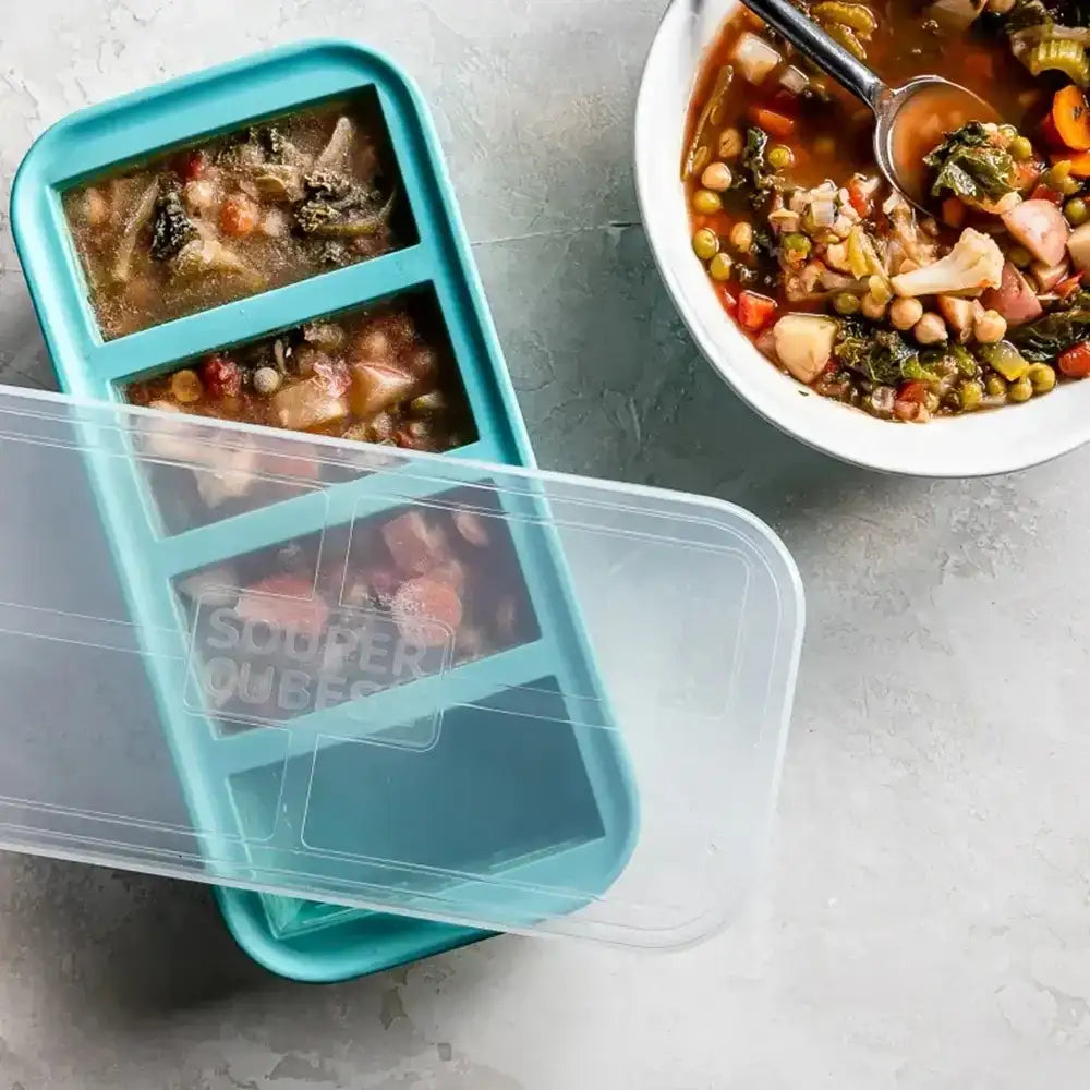 Blue freezer container with pre-made soup portions on a gray surface, next to a bowl of soup.