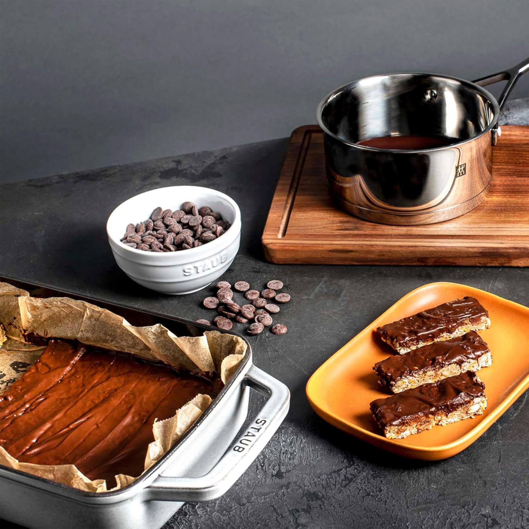 Chocolate dessert setup with a baking dish, chocolate bars, and a bowl of chocolate chips on a dark surface.