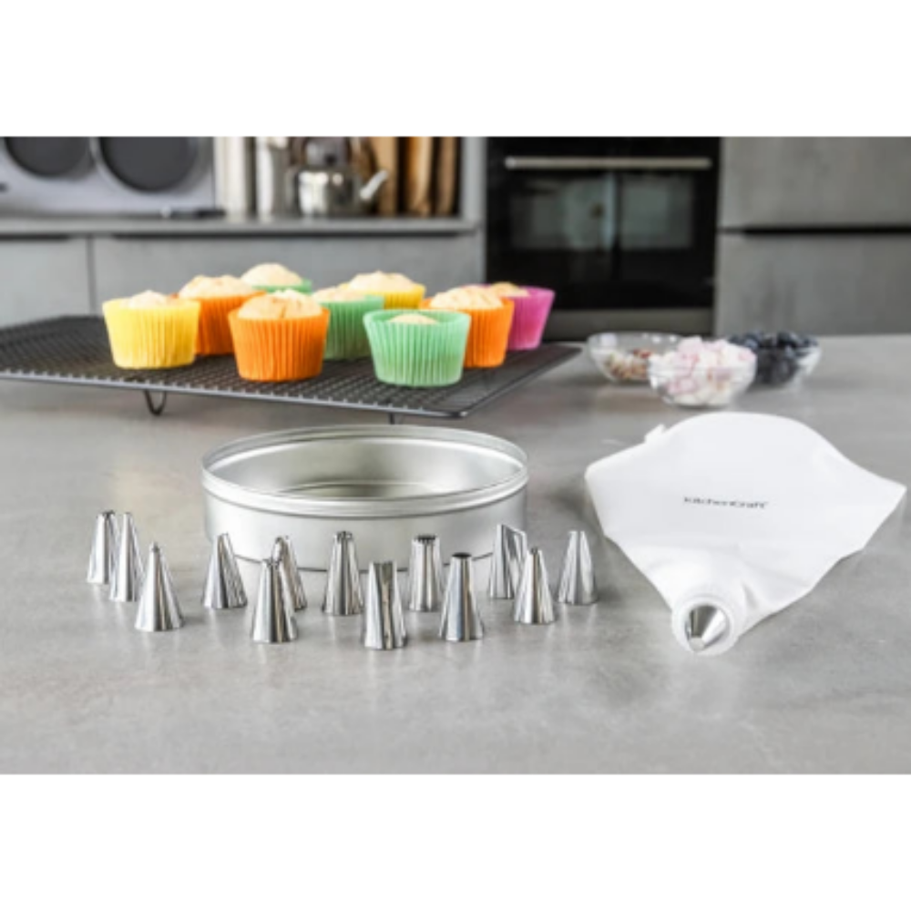 A baking accessory set on a gray kitchen countertop, including a round metal pan, multiple silver piping nozzles, and a white piping bag with the KitchenCraft logo. In the background, colorful cupcakes sit on a cooling rack in a modern kitchen.