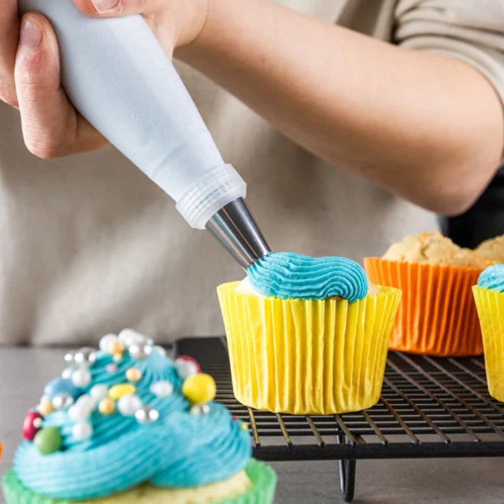 A close-up of a person piping bright blue frosting onto a cupcake in a yellow paper liner, with other decorated cupcakes on a cooling rack in the background.
