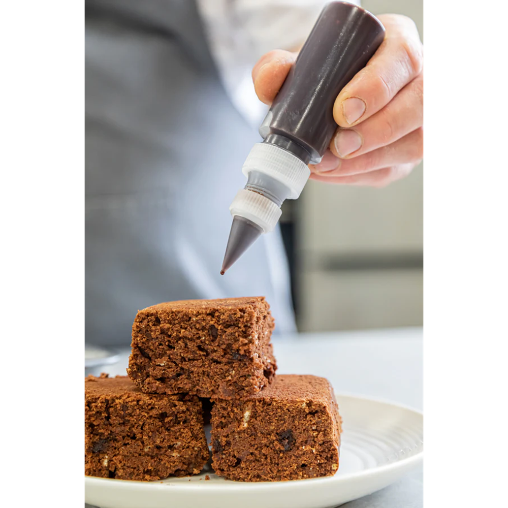 Hand using a piping bag to add chocolate frosting onto brownies on a white plate.