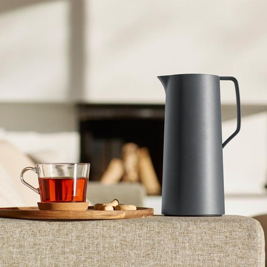 Grey pitcher on a surface with a glass of tea and snacks in the background