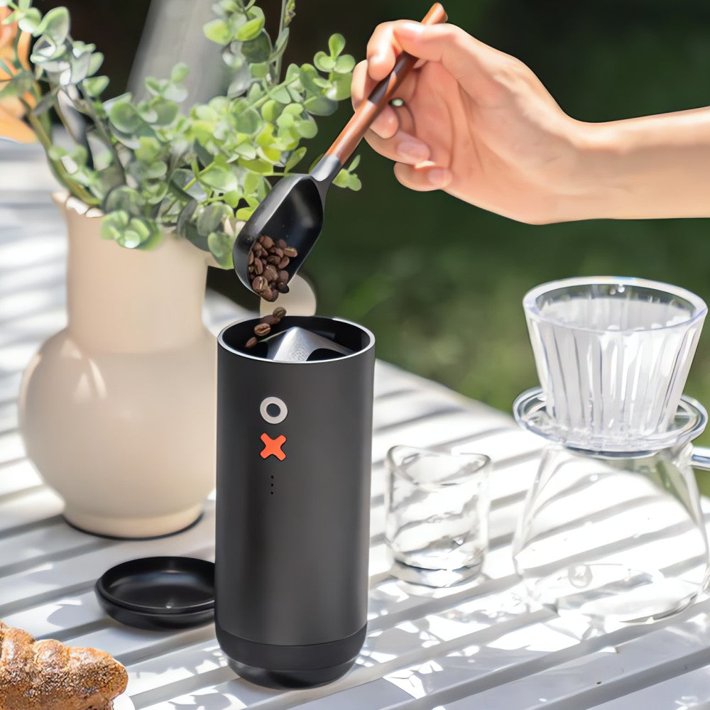 A person's hand is shown using a metal scoop to pour coffee beans into a matte black cylindrical coffee grinder. The grinder features a minimalist design with a white circular logo and an orange 'X' accent on its front.