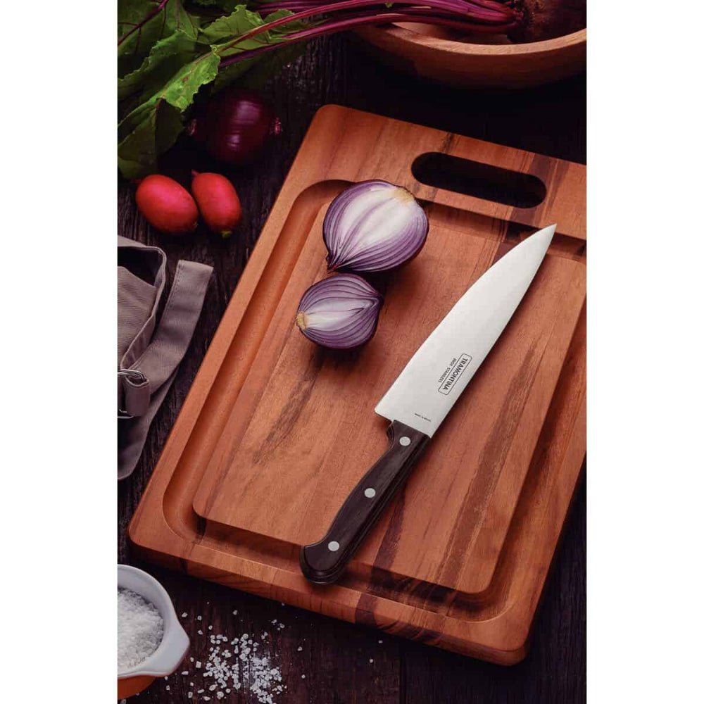 A kitchen knife with a silver blade and black handle resting on a wooden cutting board, alongside a halved red onion, radishes, and leafy greens on a dark rustic table.