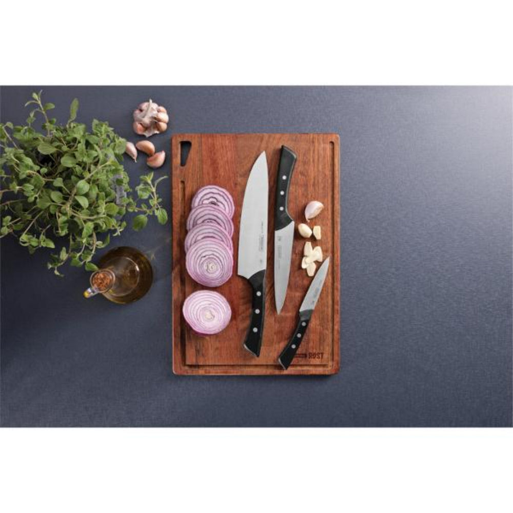 Three knives on a wooden cutting board with sliced onion and garlic, placed on a dark surface.