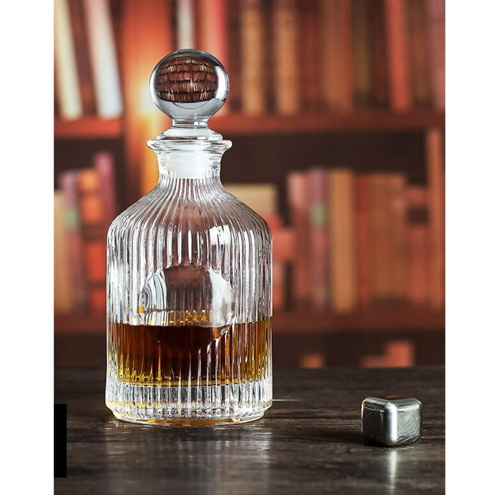 Glass whiskey decanter with wooden stopper on a wooden surface, with books in the background.