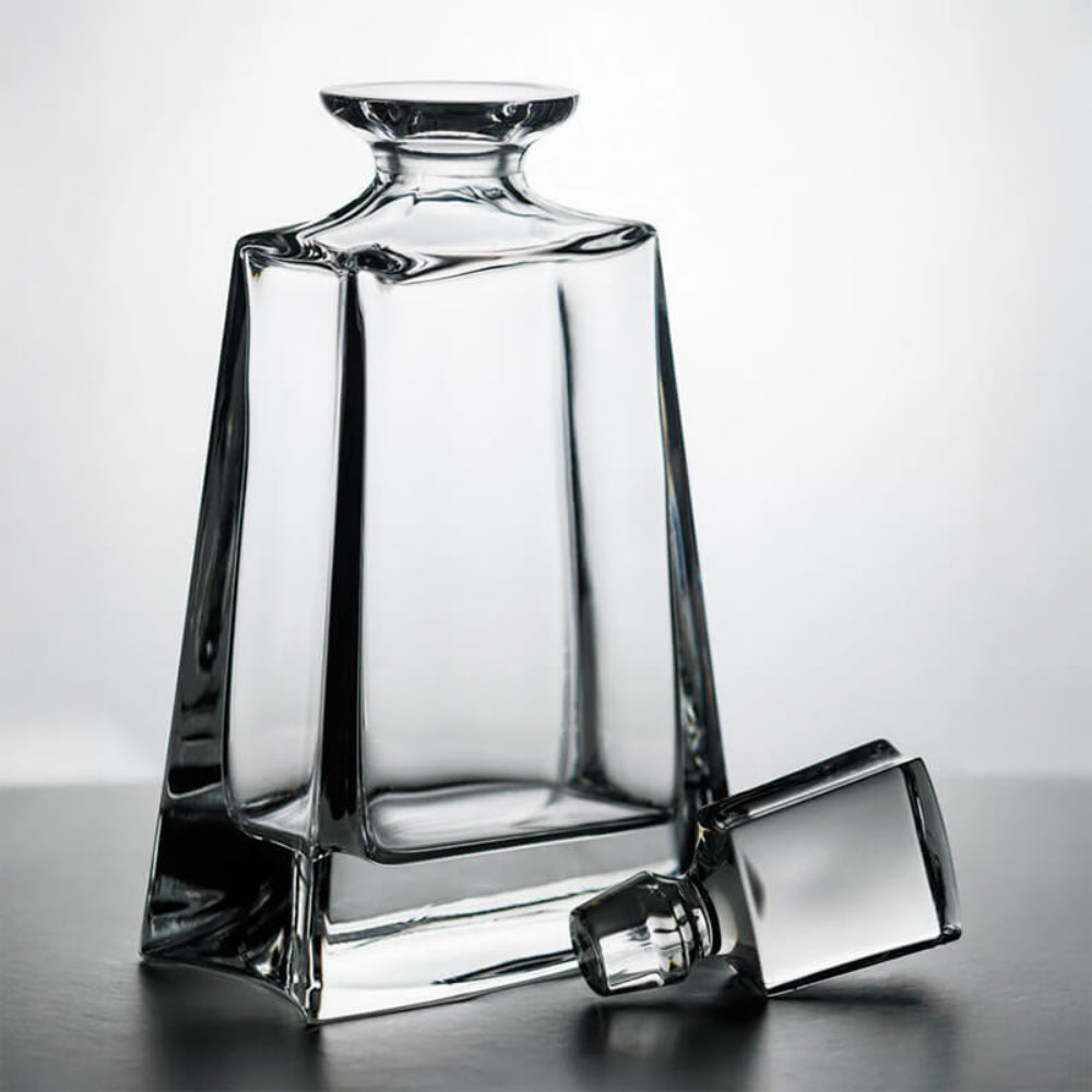 A clear glass decanter with a matching small cap placed beside it on a dark surface against a light gray background.