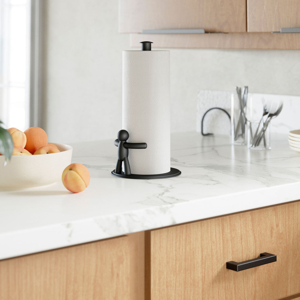 A countertop paper towel holder with a white paper towel roll, shown in a bright kitchen setting with wood cabinets, a marble counter, and fruit in a bowl.