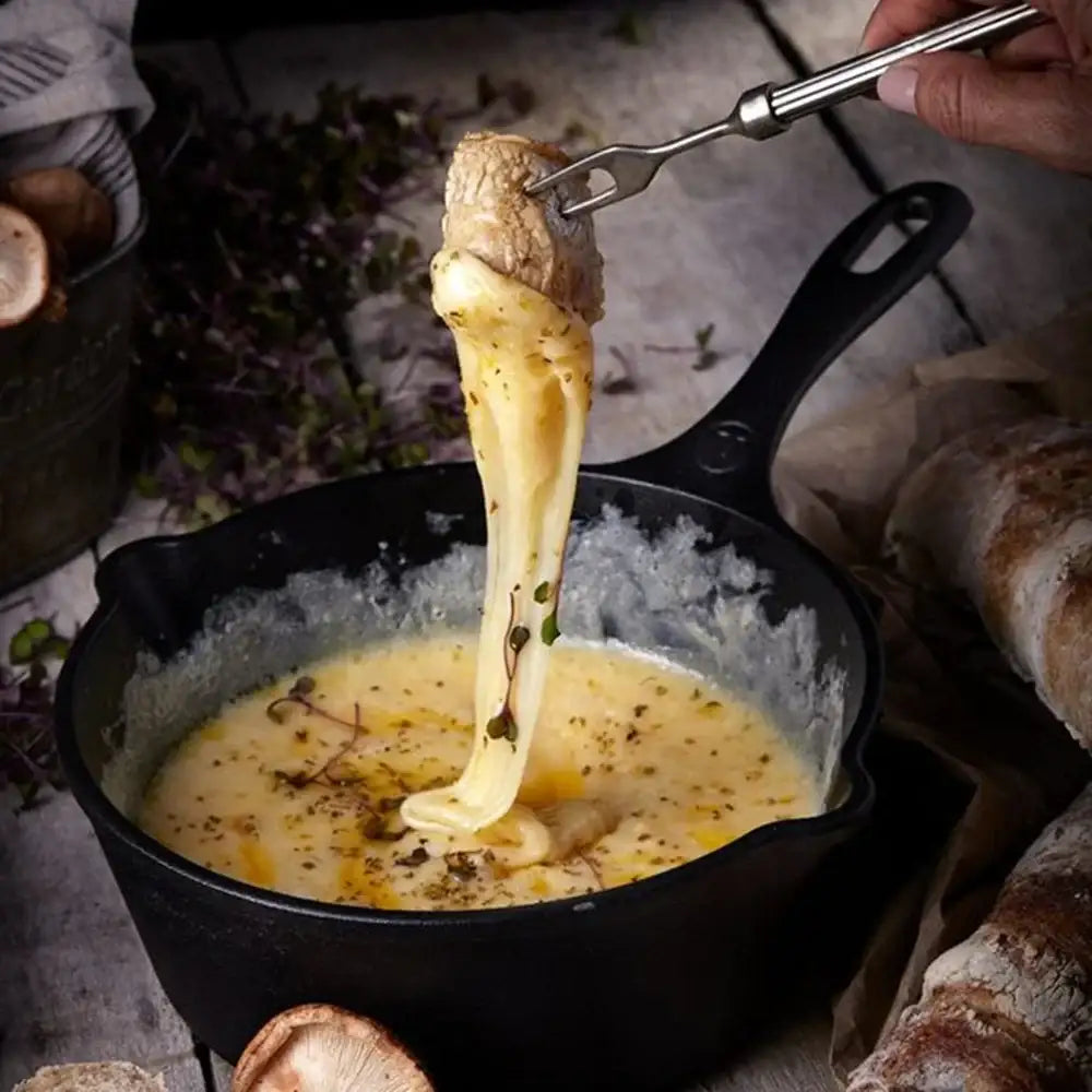 Cheese fondue being dipped into a black skillet with bread and mushrooms around.