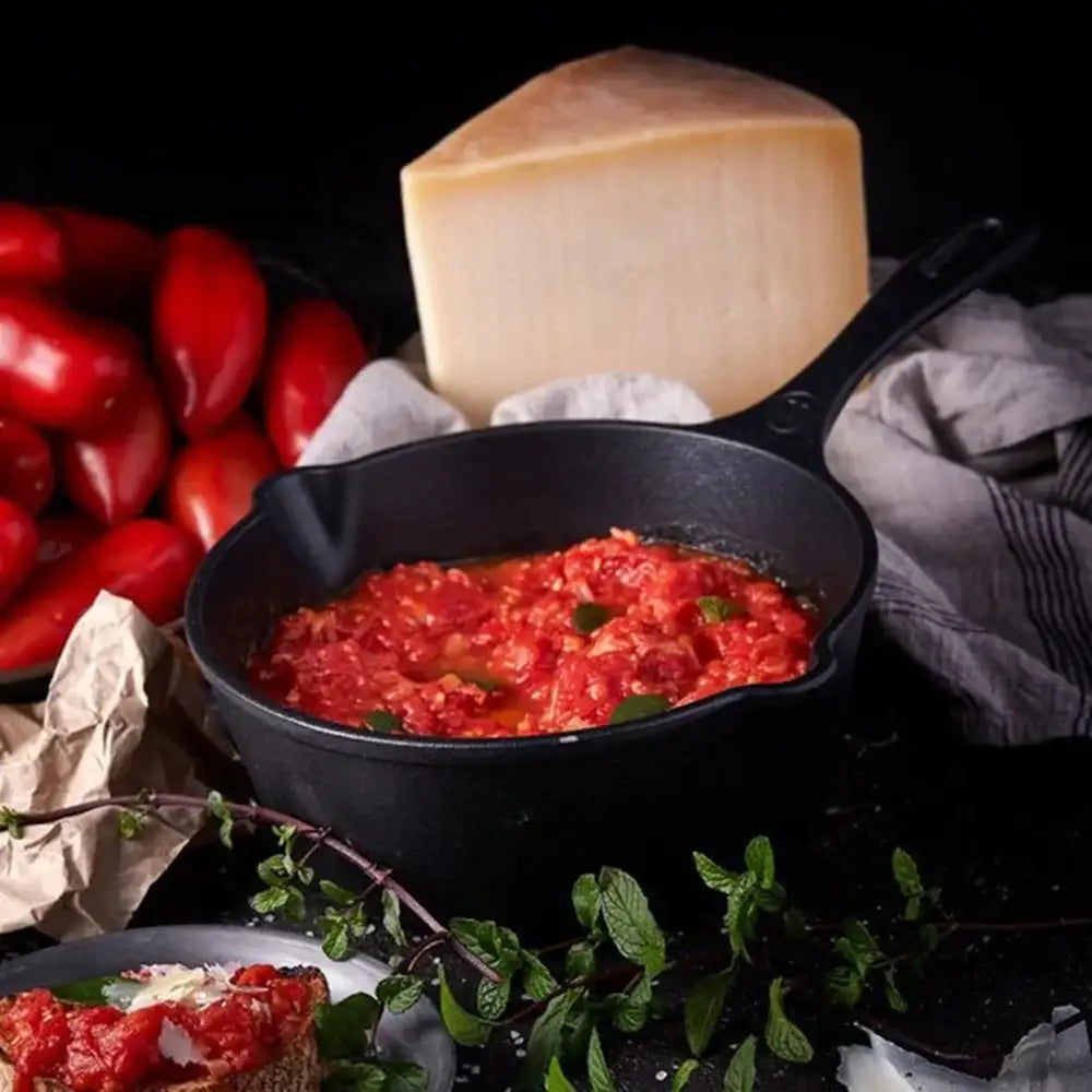 Black cast iron skillet with red tomato-based dish, surrounded by tomatoes and cheese on a dark background
