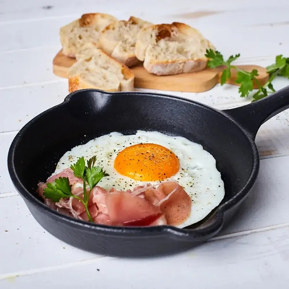 Fried egg with ham in a black skillet on a wooden surface with bread in the background
