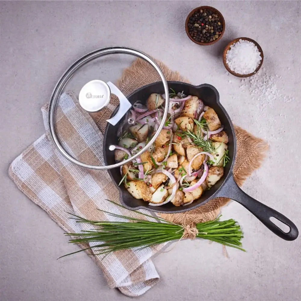 Black cast iron skillet with food on a gray surface, surrounded by herbs and small bowls of spices.