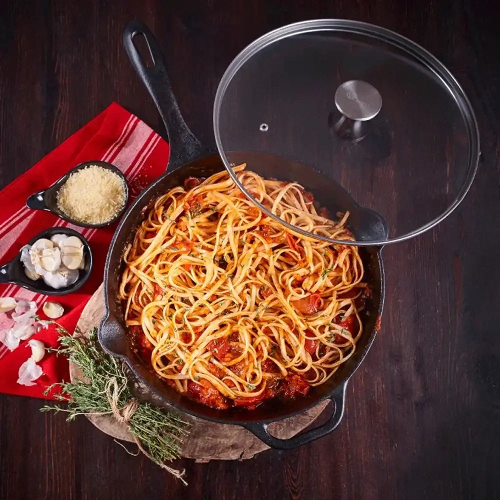 Skillet of spaghetti with a glass lid on a wooden surface
