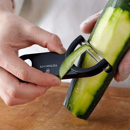Person using a Victorinox vegetable peeler to slice a zucchini on a wooden surface.