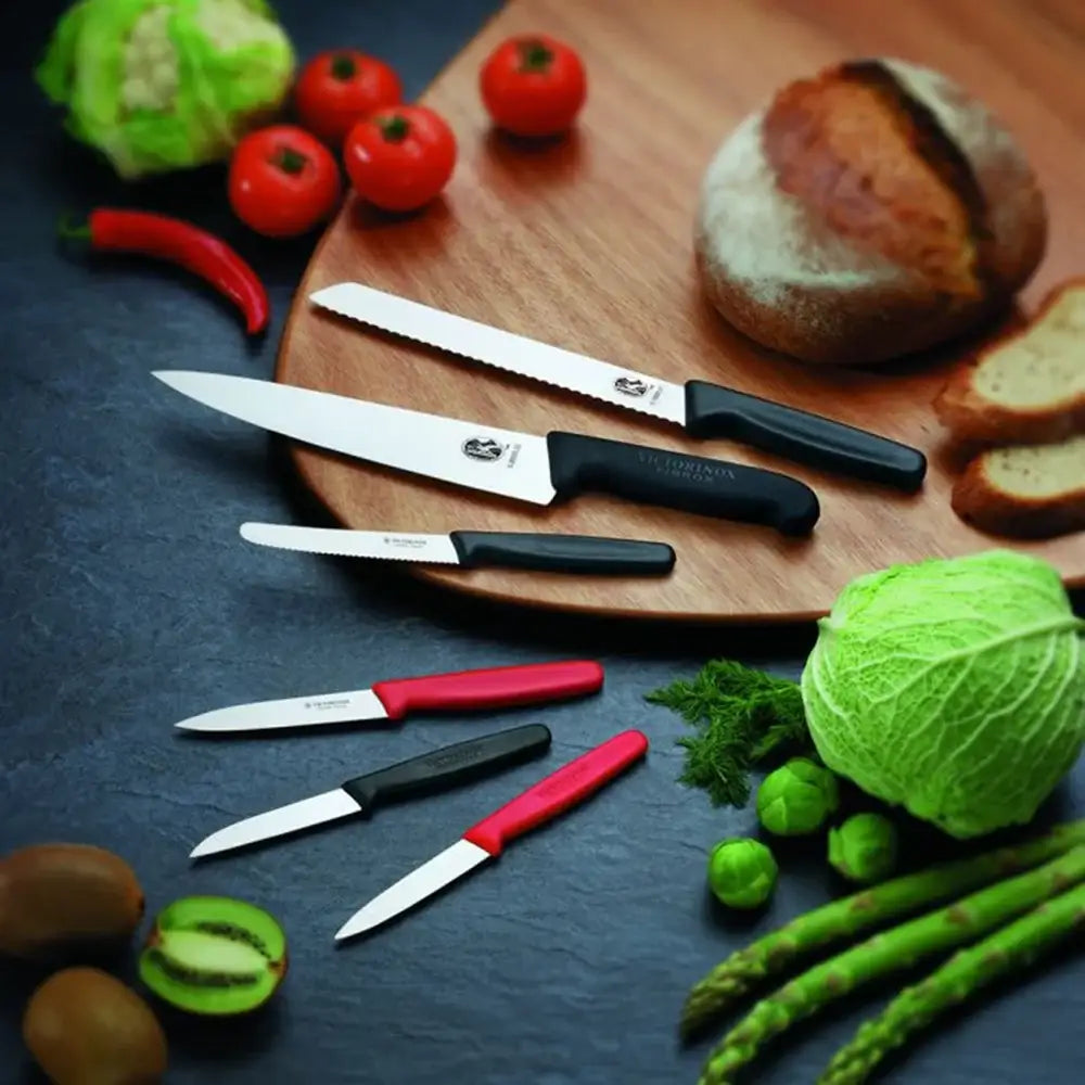 Set of knives with black and red handles on a wooden cutting board surrounded by vegetables and bread.