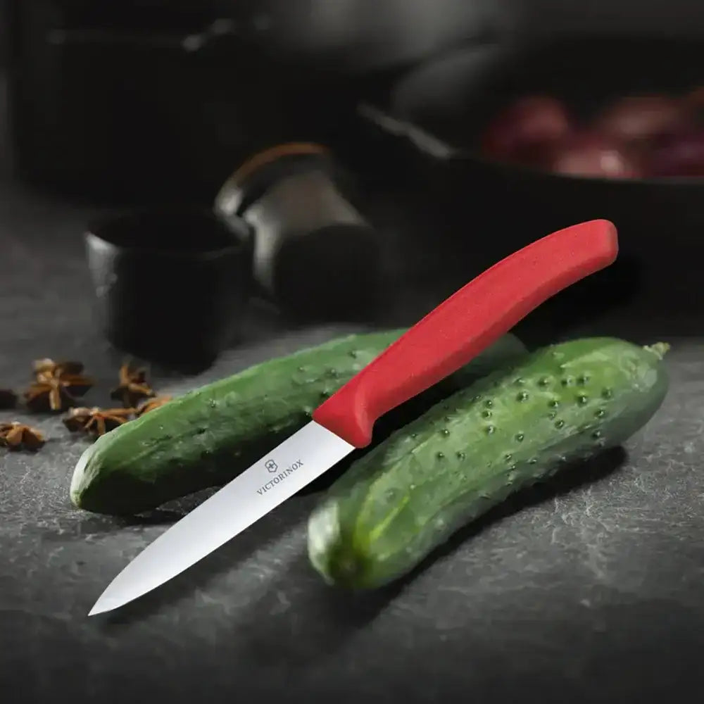 Paring knife with red handle on a cucumber on a dark surface