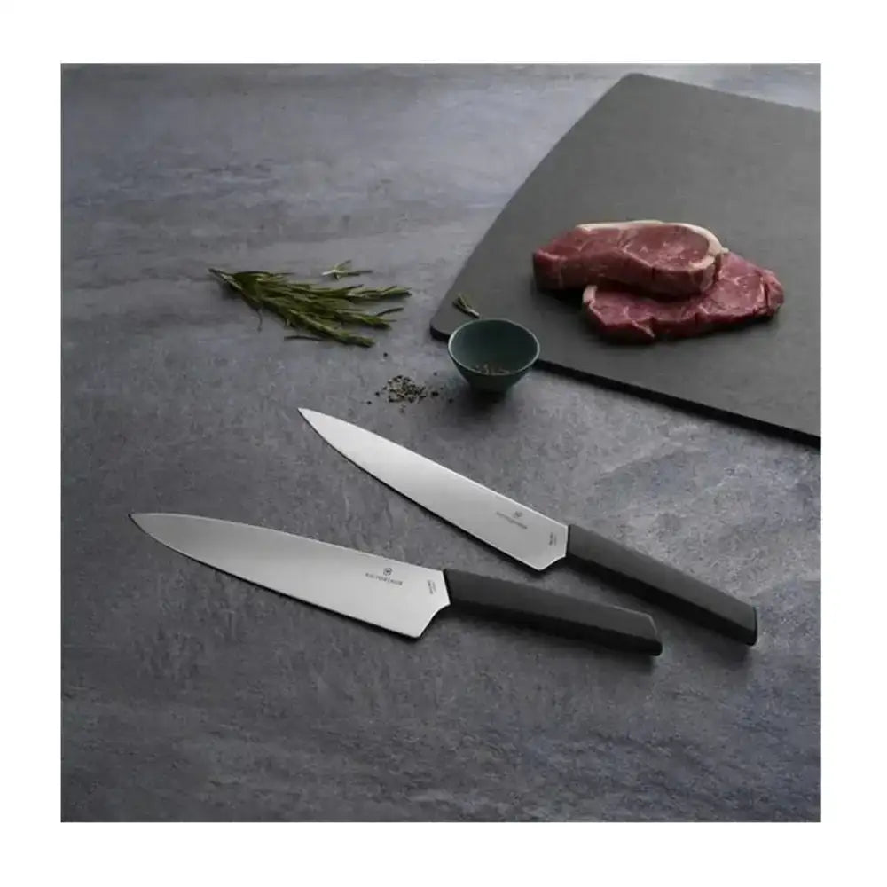 Two knives on a dark surface with a steak and herbs in the background
