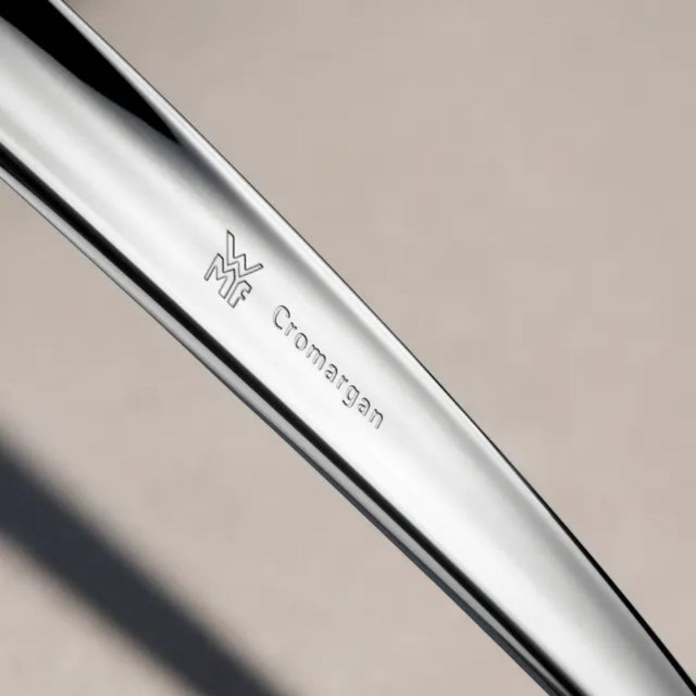 Close-up of a polished metallic silver knife blade blank with the engraved Manfredis 'Mf' logo and 'Cromargan' text on a warm beige background.