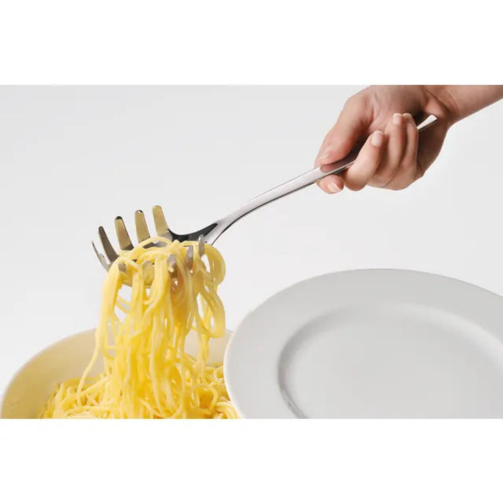 A hand holding a stainless steel spaghetti fork, lifting yellow pasta from a bowl, with a white plate in the foreground on a white background.