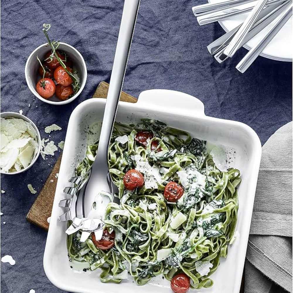 A white rectangular ceramic pasta dish with handles, filled with green tagliatelle pasta, cherry tomatoes, and grated cheese, with a large metal serving spoon resting inside. The dish is set on a dark textured tablecloth with small bowls of ingredients and silver cutlery nearby.