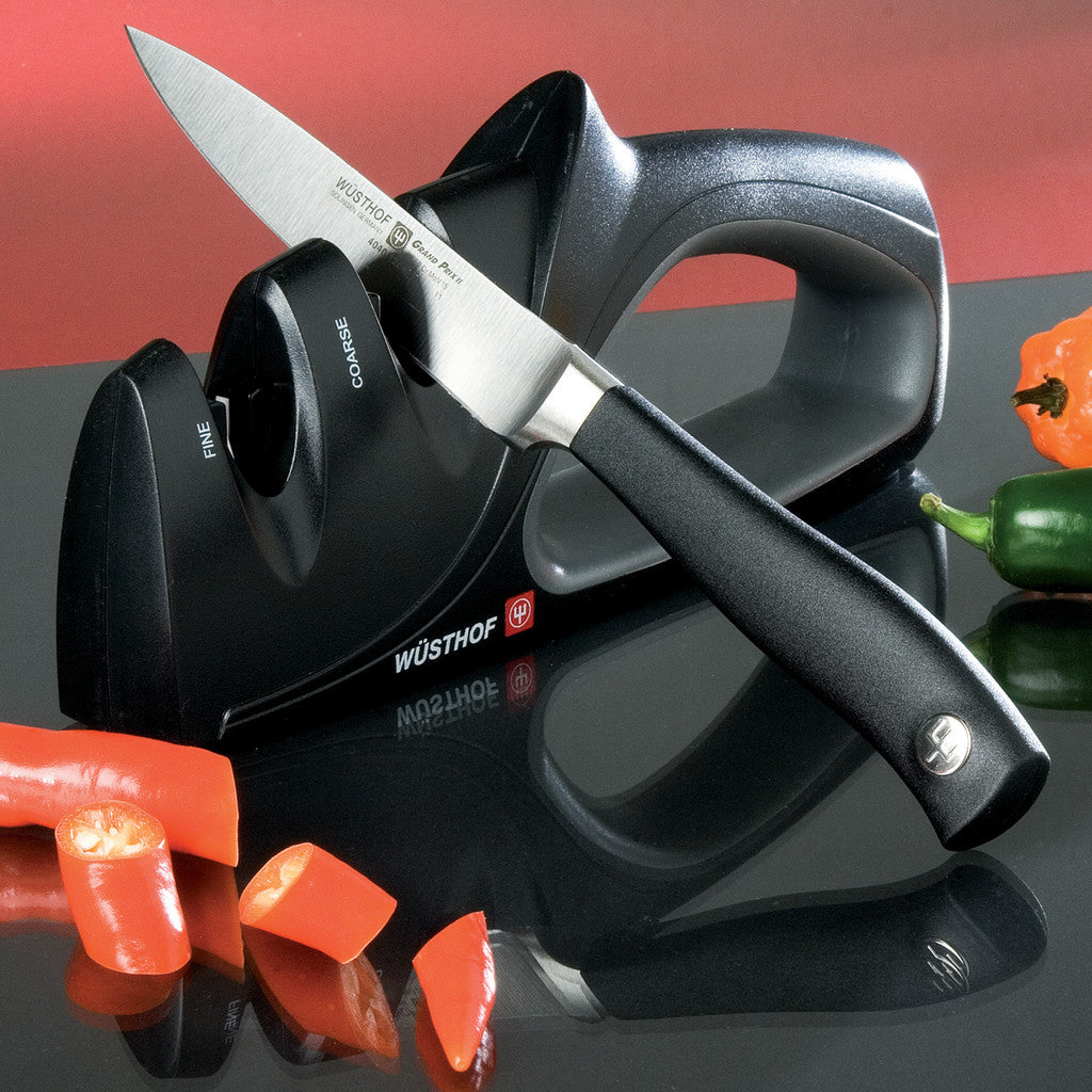 A Wusthof knife sharpening system shown with a sharpening stone and a knife, surrounded by sliced red and green peppers on a reflective black surface against a red and black background.