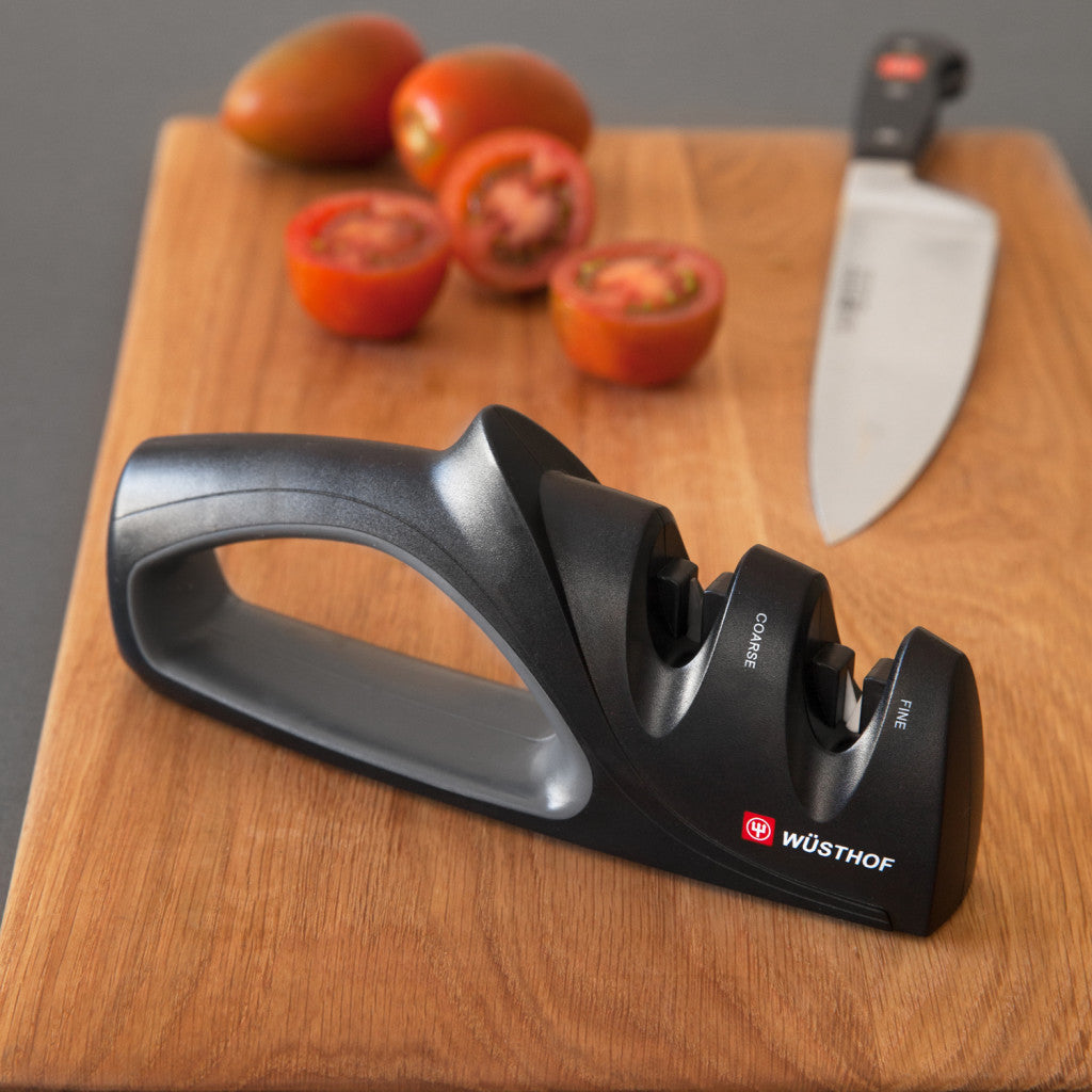 A black Wüsthof sharpening steel with a handle, placed on a wooden cutting board next to a chef's knife and sliced tomatoes.
